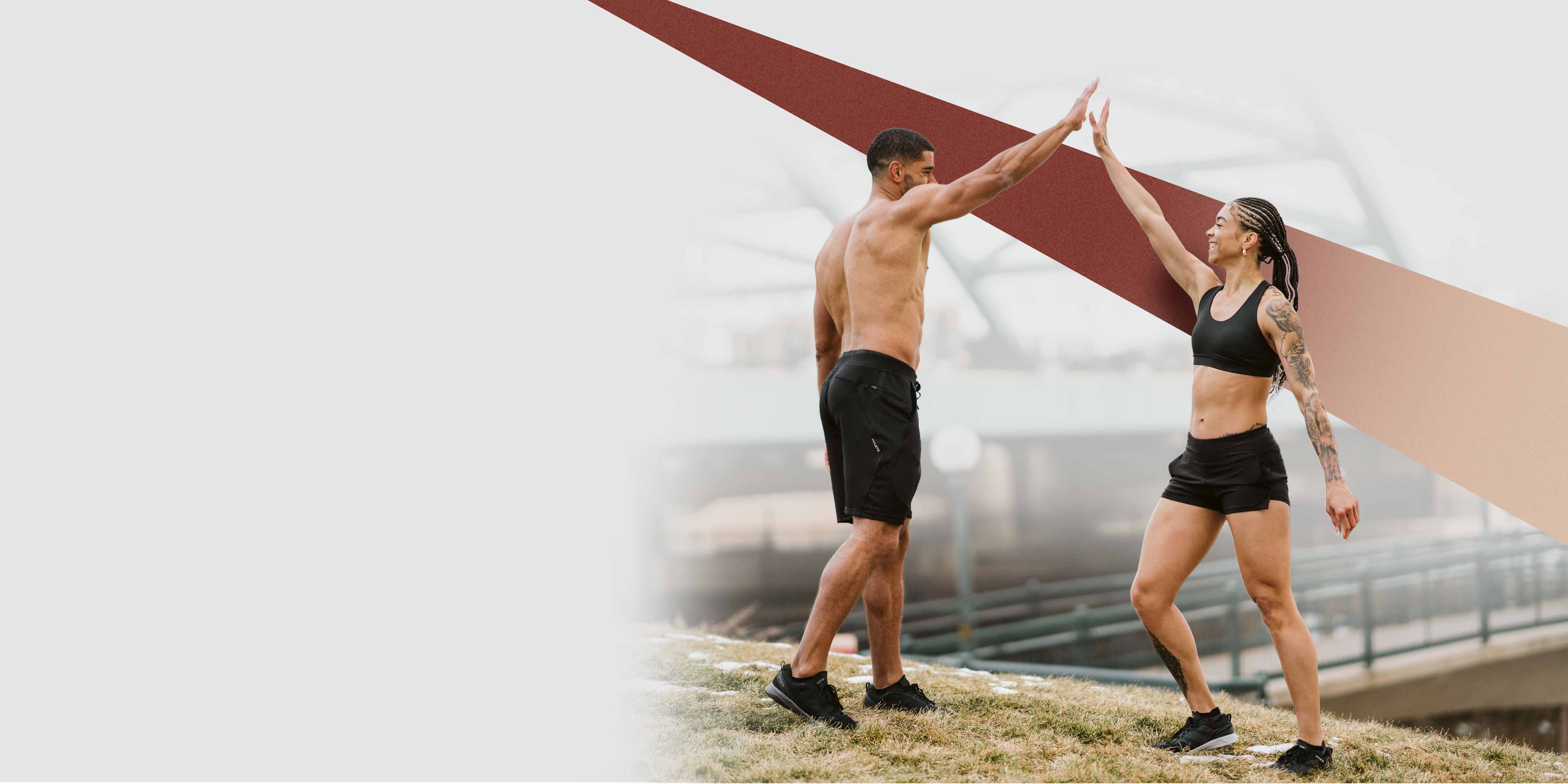 A muscular, shirtless man and a woman with an arm tattoo, both wearing Hylete black shorts, enthusiastically high-five outdoors on a grassy hill in a cool-weather setting.